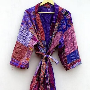 Könnte beinhalten: Ein langärmeliger Morgenmantel mit Patchwork-Design aus verschiedenen Stoffbahnen in Lila-, Rosa- und Orangetönen. Das Kleidungsstück ist mit sichtbaren weißen Steppstichdetails versehen und enthält einen passenden Bindegürtel aus Stoff.