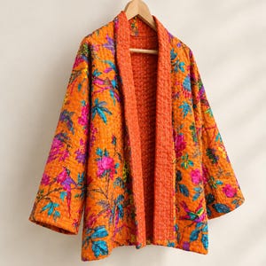 Pode incluir: Um casaco acolchoado laranja com um padrão vibrante de flores e pássaros em tons de rosa, azul e verde. A peça tem mangas compridas, uma gola larga e costuras brancas visíveis em todo o tecido, pendurada num cabide de madeira.