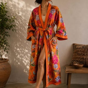 May include: A long-sleeved, floor-length robe in a vibrant orange hue, featuring a multi-coloured floral pattern in shades of pink, purple, and green. The garment includes a matching tie belt at the waist, wide sleeves, and two front pockets.
