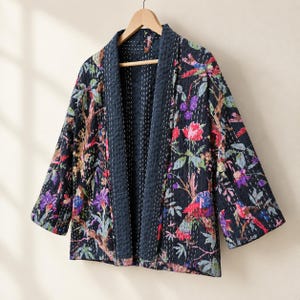 May include: A dark navy blue quilted jacket featuring a vibrant floral and bird print with pink, purple, and green accents. The garment has a solid navy blue collar and cuffs, long sleeves, and visible white kantha-style running stitch detailing.