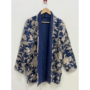Peut inclure: Une veste matelassée à manches longues présentant une base bleu foncé avec un imprimé floral et botanique beige clair. Le vêtement possède une doublure intérieure et une patte de boutonnage bleu foncé unies, rehaussées de surpiqûres blanches.