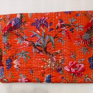 Puede incluir: Una colcha rectangular de color naranja con un patrón repetitivo de pájaros coloridos, grandes flores moradas y rosas, y follaje verde. La tela presenta bordados visibles de puntada recta en color blanco por toda la superficie.