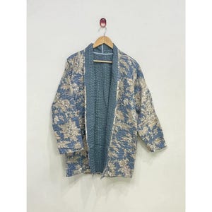 Peut inclure: Une veste matelassée présentant un motif floral bleu et beige avec des détails de points de couture blancs visibles. Le vêtement possède des manches longues, une ouverture sur le devant et une doublure intérieure bleue unie, sur un cintre.