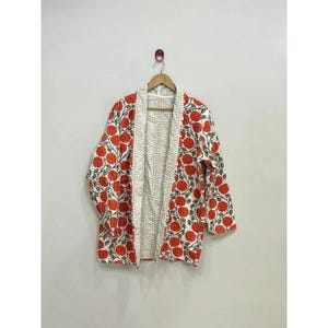 Peut inclure: Une veste à manches longues ouverte sur le devant, présentant une base blanche avec un motif répétitif de fleurs orange et de feuilles vertes. Le vêtement possède une texture matelassée et est présenté sur un cintre en bois.