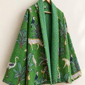 Pode incluir: Um casaco acolchoado verde com uma estampa vibrante de selva com leopardos, tucanos e palmeiras. A peça possui gola larga, mangas compridas e costuras brancas visíveis por todo o tecido, pendurada em um cabide de madeira.