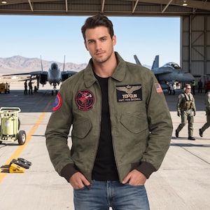 Könnte beinhalten: Eine olivgrüne Bomberjacke über einem schwarzen T-Shirt und blauen Jeans, verziert mit verschiedenen gestickten Aufnähern, darunter ein Top Gun-Logo mit dem Text Capt Pete Mitchell, ein kreisförmiges rotes Teufels-Emblem und eine kleine US-Flagge.