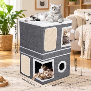 May include: A two-story gray fabric cat condo with white plush trim, featuring integrated sisal scratching pads, circular cutouts, and hanging toy balls. The structure measures 16.14 inches wide, 16.14 inches deep, and 32.68 inches in total height.