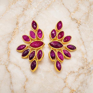 May include: A pair of gold-toned earrings featuring a cluster of faceted, marquise-cut magenta stones arranged in a fan-like, leaf-inspired pattern. Each earring consists of multiple individual stones set in a gold metal frame, creating a symmetrical design that radiates outward from a central, larger marquise-shaped stone.
