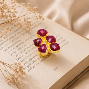 Könnte beinhalten: Ein goldfarbener Ring mit fünf facettierten, tief rosa-roten Steinen, die in einer Gruppe angeordnet sind. Die Steine haben eine abgerundete, unregelmäßige Form und sind in ein goldenes Metallband gefasst, das die blütenartige Anordnung trägt.