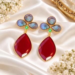 Può includere: Un paio di orecchini pendenti dorati con tre pietre di labradorite sfaccettate e iridescenti a forma di trifoglio, arricchite da una piccola gemma verde e una grande pietra rosso scuro sfaccettata a forma di goccia.