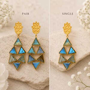 Puede incluir: Pendientes colgantes dorados con una pieza superior en forma de mano de Hamsa texturizada y tres niveles de piedras triangulares de labradorita azul iridiscente. La imagen muestra un par a la izquierda y un pendiente a la derecha, marcados PAIR y SINGLE.
