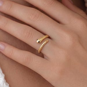 Könnte beinhalten: Ein goldfarbener Ring in Form einer gewundenen Schlange mit einem strukturierten, schuppenartigen Muster entlang der Ringschiene. Der Ring hat einen glatten, abgerundeten Kopf und ein spitz zulaufendes Ende, das sich offen um den Finger legt.
