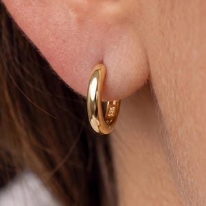 Può includere: Un piccolo orecchino a cerchio dorato lucido con una superficie liscia e arrotondata. L'orecchino presenta un marchio 925 visibile sul lato interno del cerchio, che indica una base in argento sterling con finitura dorata, indossato al lobo.