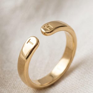 Puede incluir: Un anillo abierto de tono dorado con un acabado liso y pulido. Los dos extremos de la banda son redondeados y cada uno está grabado con una letra mayúscula, específicamente la letra T en un lado y la letra H en el otro lado.