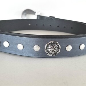 Op de afbeelding: Een grijze leren riem met zilverkleurige metalen accenten. De riem heeft een decoratief bloemmotief op de gesp en verschillende kleinere ronde accenten langs de band.