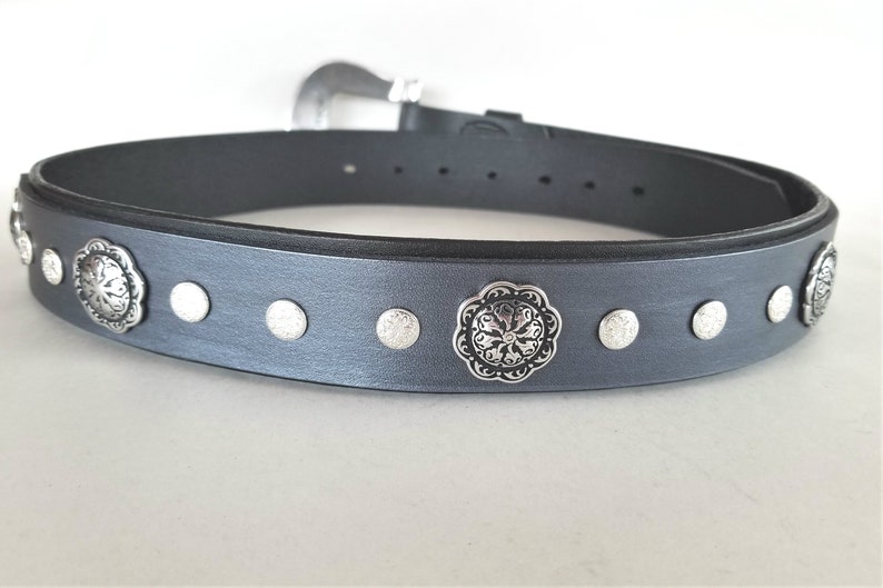 Op de afbeelding: Een grijze leren riem met zilverkleurige metalen accenten. De riem heeft een decoratief bloemmotief op de gesp en verschillende kleinere ronde accenten langs de band.