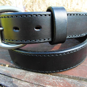 May include: A black leather belt with a silver buckle and double stitching. The belt is folded in half and lying on a wooden surface.