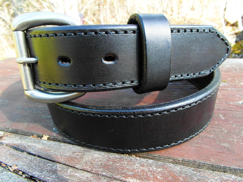 May include: A black leather belt with a silver buckle and double stitching. The belt is folded in half and lying on a wooden surface.