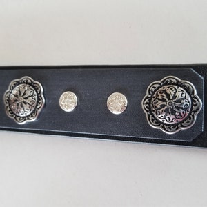 Op de afbeelding: Zwarte leren riem met zilverkleurige decoratieve accenten. De riem heeft een bloemmotief en kleine ronde studs.