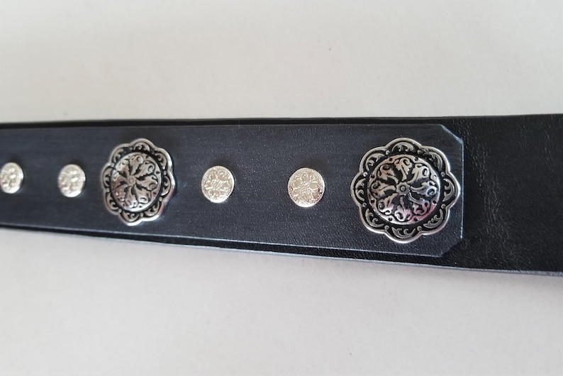 Op de afbeelding: Zwarte leren riem met zilverkleurige decoratieve accenten. De riem heeft een bloemmotief en kleine ronde studs.