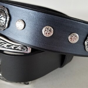 Op de afbeelding: Een zwarte leren riem met een zilveren gesp en decoratieve zilveren accenten. De riem heeft een bloemmotief op de gesp en accenten.