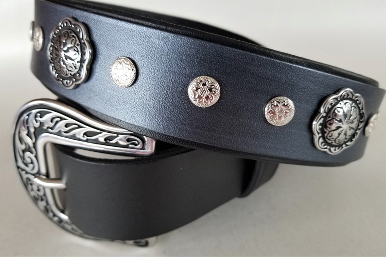 Op de afbeelding: Een zwarte leren riem met een zilveren gesp en decoratieve zilveren accenten. De riem heeft een bloemmotief op de gesp en accenten.