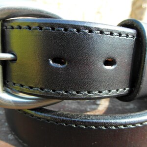 May include: A black leather belt with a silver buckle and stitching. The belt has two rows of stitching and two holes for adjusting the size.