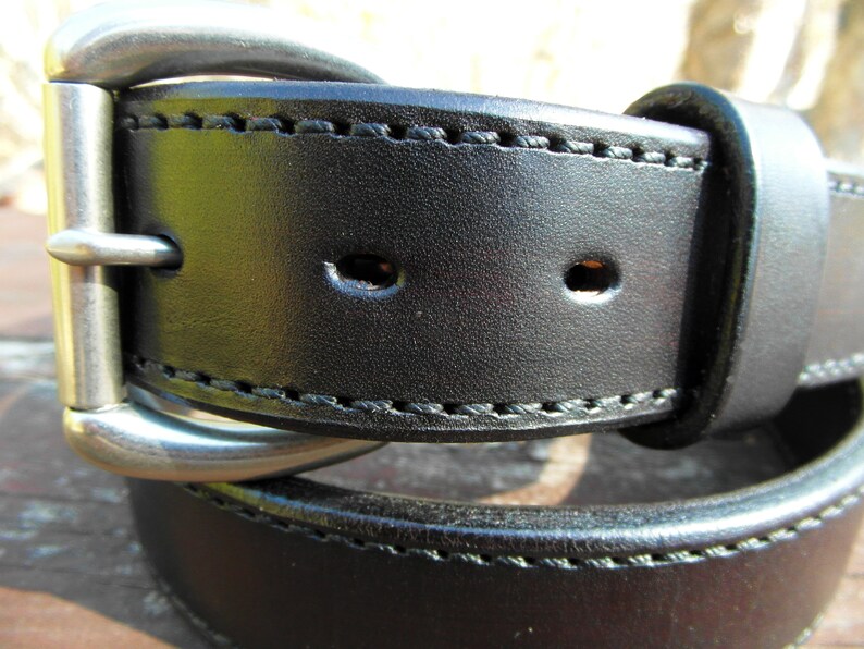 May include: A black leather belt with a silver buckle and stitching. The belt has two rows of stitching and two holes for adjusting the size.