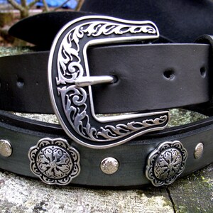 Op de afbeelding: Een zwarte leren riem met een zilveren gesp en decoratieve metalen accenten. De gesp heeft een bloemmotief.