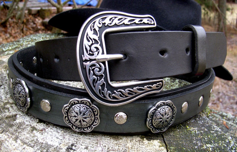 Op de afbeelding: Een zwarte leren riem met een zilveren gesp en decoratieve metalen accenten. De gesp heeft een bloemmotief.