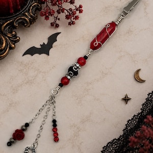 May include: A metal roach clip featuring a red cylindrical stone wrapped in silver wire, accented with black and red beads. The end has three silver chains dangling a red rose charm, a silver vampire fang charm, and small black and red crystal beads.