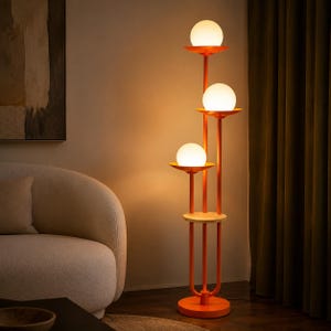 May include: A tall floor lamp featuring a vibrant orange metal frame with three vertical poles of varying heights. Each pole is topped with a circular orange tray holding a glowing white spherical glass globe light. A small circular wooden shelf is attached.