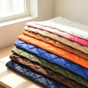 May include: A stack of folded quilted fabric pieces in various colours including white, pink, tan, orange, blue, olive green, brown, and dark navy. Each piece features a consistent diamond-shaped stitched pattern across the entire surface area.