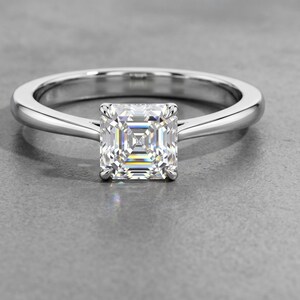 May include: A silver-coloured metal ring featuring a single square-cut clear gemstone set in a four-prong setting. The band is smooth and polished, tapering slightly as it connects to the central stone, creating a clean and reflective appearance.
