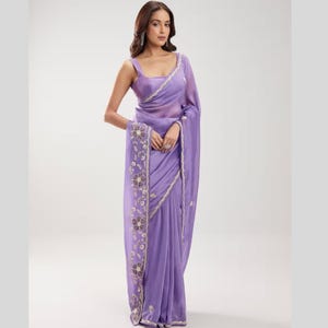 May include: A lavender saree featuring a sleeveless blouse and a matching drape. The fabric has a smooth, satin-like finish, and the border is decorated with intricate floral embroidery in silver and dark thread, creating a detailed pattern.