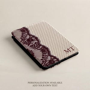 May include: A rectangular electronic device cover featuring a cream-colored background with small black polka dots. A dark burgundy lace pattern runs diagonally across the left side. The letters MT are printed in dark burgundy on the right side.