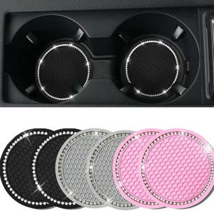 May include: A set of six circular car cup holder inserts featuring a textured honeycomb pattern and a border of embedded clear rhinestones. The set includes two black, two gray, and two pink silicone coasters designed for vehicle consoles.