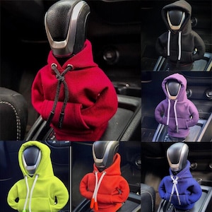 May include: A collection of miniature fabric hoodies designed to fit over a car gear shift knob. The hoodies are available in various solid colours including red, black, purple, neon yellow, orange, and blue, each featuring a hood and drawstrings.