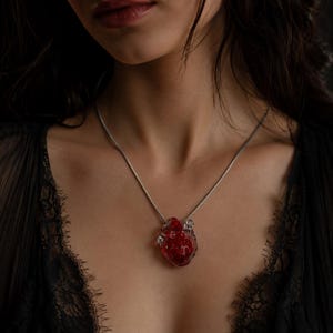 Puede incluir: Un collar con un colgante en forma de coraz&oacute;n humano anat&oacute;mico realista. El coraz&oacute;n est&aacute; hecho de material rojo transl&uacute;cido con detalles internos sutiles. Cuelga de una cadena fina de metal de tono plateado que descansa sobre el cuello.