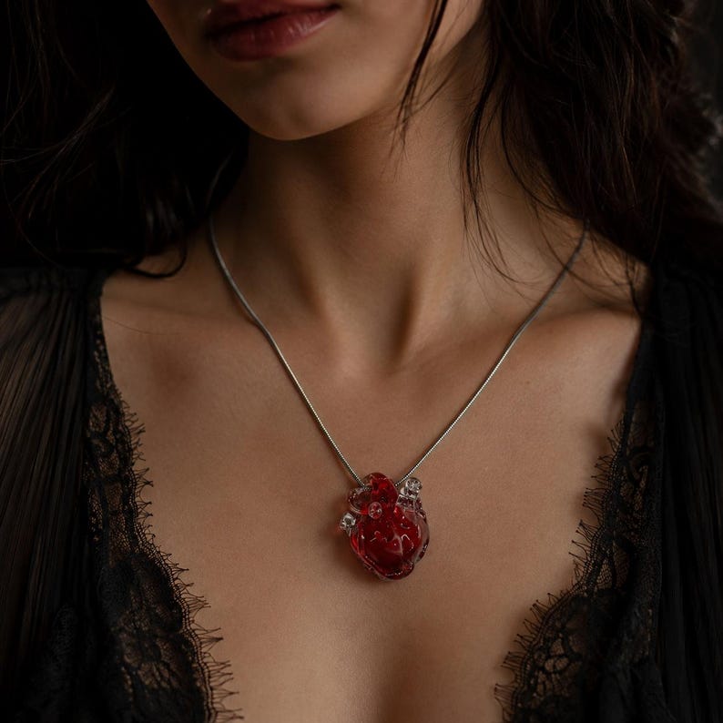 Puede incluir: Un collar con un colgante en forma de coraz&oacute;n humano anat&oacute;mico realista. El coraz&oacute;n est&aacute; hecho de material rojo transl&uacute;cido con detalles internos sutiles. Cuelga de una cadena fina de metal de tono plateado que descansa sobre el cuello.