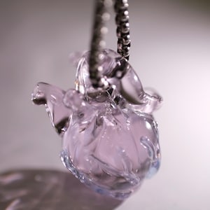 Puede incluir: Un colgante de cristal transl&uacute;cido de color morado claro con forma de coraz&oacute;n anat&oacute;mico, suspendido de una cadena de bolas de metal plateado. El cristal presenta contornos suaves y redondeados con un acabado brillante que capta la luz.