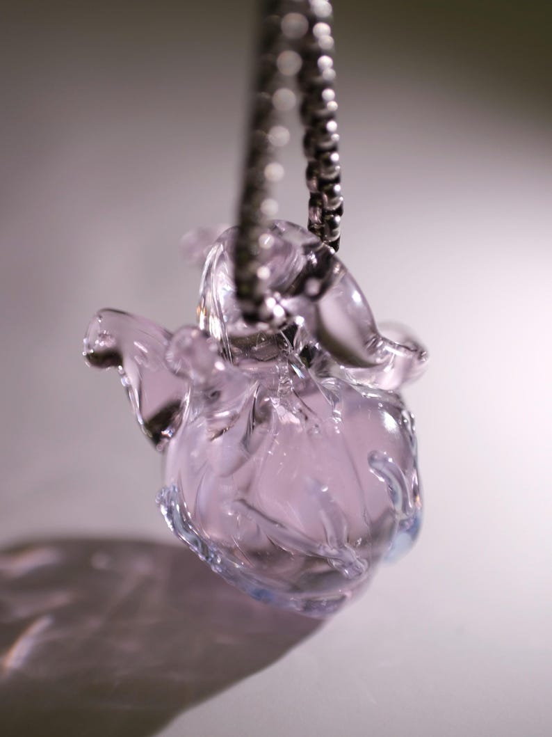 Puede incluir: Un colgante de cristal transl&uacute;cido de color morado claro con forma de coraz&oacute;n anat&oacute;mico, suspendido de una cadena de bolas de metal plateado. El cristal presenta contornos suaves y redondeados con un acabado brillante que capta la luz.