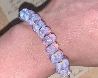 Puede incluir: Una pulsera compuesta por cuentas de vidrio redondas y translúcidas con un acabado iridiscente y opalescente que refleja la luz en tonos de azul suave, rosa y lavanda. Las cuentas están ensartadas en una sola fila, creando un efecto brillante.