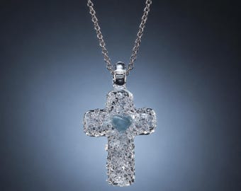 Puede incluir: Un colgante en forma de cruz con una superficie cristalina transparente y texturizada, que presenta una piedra lisa en forma de corazón de color azul claro en el centro. El colgante cuelga de una fina cadena de eslabones de metal plateado.