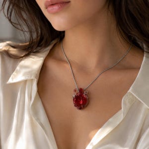 Puede incluir: Un collar con un colgante de vidrio transl&uacute;cido de color rojo intenso con forma de coraz&oacute;n anat&oacute;mico. El colgante cuelga de una cadena de metal fino y liso de tono plateado que descansa sobre la clav&iacute;cula, mostrando los detalles del vidrio.