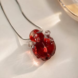 Puede incluir: Un collar con un colgante en forma de coraz&oacute;n anat&oacute;mico hecho de vidrio transl&uacute;cido de color rojo intenso. El colgante cuelga de una fina cadena de serpiente de metal plateado. El coraz&oacute;n de vidrio muestra detalles intrincados y brillos.