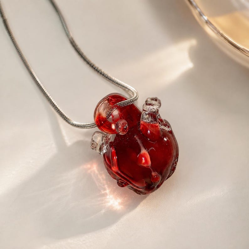 Puede incluir: Un collar con un colgante en forma de coraz&oacute;n anat&oacute;mico hecho de vidrio transl&uacute;cido de color rojo intenso. El colgante cuelga de una fina cadena de serpiente de metal plateado. El coraz&oacute;n de vidrio muestra detalles intrincados y brillos.