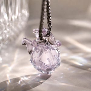 Puede incluir: Un colgante de cristal transl&uacute;cido de color violeta claro con forma de peque&ntilde;a criatura redondeada, con extremidades delicadas y curvas. El colgante cuelga de una cadena de metal oscuro, captando la luz para crear reflejos suaves y brillantes.