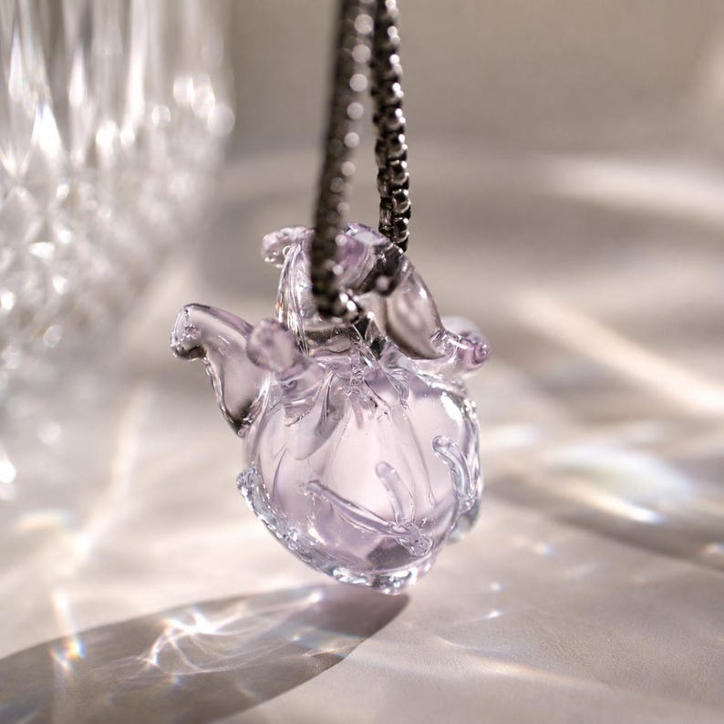 Puede incluir: Un colgante de cristal transl&uacute;cido de color violeta claro con forma de peque&ntilde;a criatura redondeada, con extremidades delicadas y curvas. El colgante cuelga de una cadena de metal oscuro, captando la luz para crear reflejos suaves y brillantes.