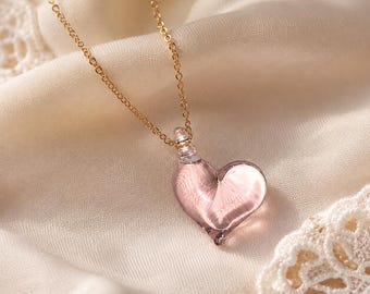 Puede incluir: Un collar de cadena en tono dorado con un colgante de corazón de vidrio translúcido en color rosa pálido. El colgante tiene forma de corazón suave y redondeado, con un asa de vidrio transparente en la parte superior que se une a la fina cadena metálica.